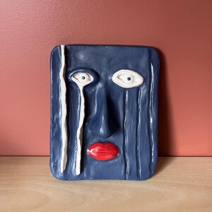 Peut inclure: Sculpture de visage en céramique bleu marine avec des yeux blancs, une bouche rouge et des traits blancs en forme de larmes. La sculpture rectangulaire a un design minimaliste et est placée sur une surface en bois.