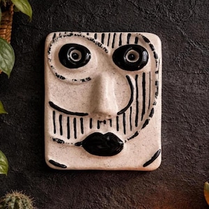May include: A rectangular ceramic wall plaque featuring a stylized face with a speckled off-white finish. The design includes two large black circular eyes, a prominent central nose, and full black lips, accented by black line details.