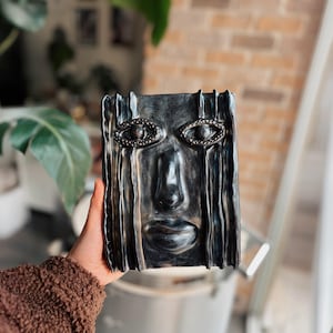 May include: A rectangular, dark gray ceramic face sculpture with detailed eyes and a defined nose and mouth. The sculpture has vertical lines that give the impression of tears. The eyes are embellished with a beaded design.