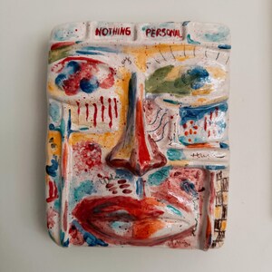May include: A rectangular ceramic wall art piece featuring a stylized face with bold colors. The face has a prominent red nose, red lips, and blue and yellow accents. The text "NOTHING PERSONAL" is written across the top.