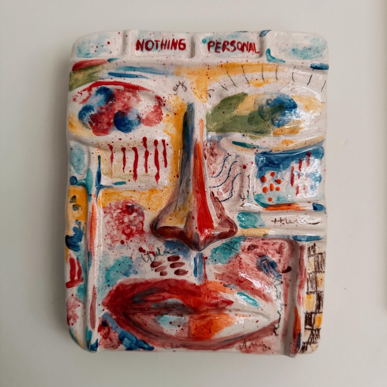 May include: A rectangular ceramic wall art piece featuring a stylized face with bold colors. The face has a prominent red nose, red lips, and blue and yellow accents. The text "NOTHING PERSONAL" is written across the top.