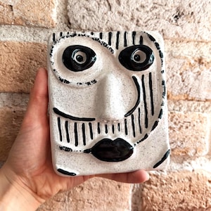 May include: A rectangular ceramic wall plaque featuring a stylised face with a speckled white glaze and bold black painted details. The design includes two large circular eyes, a prominent central nose, and a dark mouth with vertical line accents.