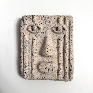 Op de afbeelding: Een rechthoekige wandplaat met een gebeeldhouwd gezicht in een gespikkelde beige en zwarte afwerking. Het ontwerp bevat verhoogde ogen, een prominente neus en een afgeronde mond, met verticale gegroefde lijnen over het oppervlak van het stuk.
