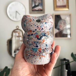 Op de afbeelding: Een gespikkelde keramische vaas in een gestapeld ontwerp, vastgehouden in een hand. De vaas is voornamelijk wit met kleurrijke verfspatten in blauw, rood, oranje en groen. De vaas heeft een kleine opening aan de bovenkant.