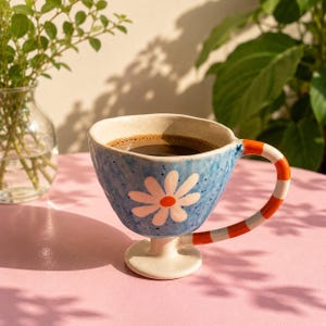 Op de afbeelding: Een keramische mok met een gespikkeld lichtblauw glazuur, voorzien van een witte margriet met een oranje hart aan de voorzijde. De mok heeft een voetstuk en een gebogen handvat met oranje en witte strepen, gevuld met donkerbruine koffie.