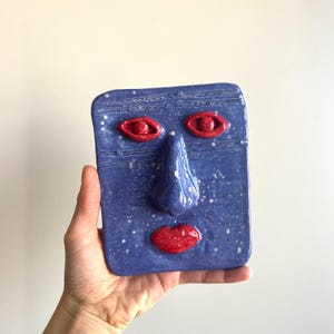 Op de afbeelding: Een blauwe keramische gezichtssculptuur met rode ogen en lippen en een prominente neus. Het vierkante stuk heeft een gespikkelde textuur en wordt in een hand gehouden. Het kunstwerk is een uniek decoratief item.
