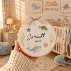 Peut inclure: Broderie sur cercle avec un thème nautique. Le design comprend une baleine, une tortue, une étoile de mer et le nom "Jarrett" avec une date. Le cercle est orné d'un nœud beige.