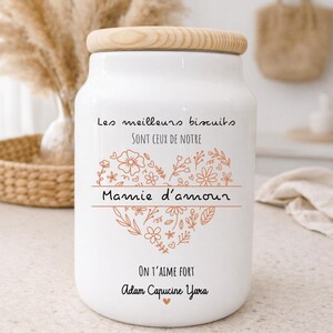 Peut inclure: Un pot à biscuits en céramique blanche avec un couvercle en bois. Le pot présente un motif floral en forme de cœur et du texte en français. Le texte comprend "Les meilleurs biscuits sont ceux de notre Mamie d'amour" et "ON T'AIME FORT Adam Capucine Yara."