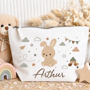 Peut inclure: Une trousse de maquillage en toile blanche avec un lapin de dessin animé, des nuages, des étoiles et un avion en papier. Le nom "Arthur" est écrit en script marron. La trousse a une fermeture éclair et est entourée de jouets pour enfants.