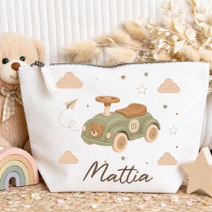 Peut inclure: Trousse de maquillage en toile blanche avec fermeture éclair. Elle présente une illustration de voiture verte avec un visage d'ours en peluche, des nuages, des étoiles et le nom "Mattia" en écriture brune. Un joli cadeau pour bébé.