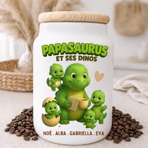 Peut inclure: Pot en céramique blanc avec un couvercle en bois, orné d'une illustration de famille de dinosaures verts. Le texte "PAPASAURUS ET SES DINOS" est affiché au-dessus de la famille. Les noms NOÉ, ALBA, GABRIELLA et EVA sont imprimés en bas.