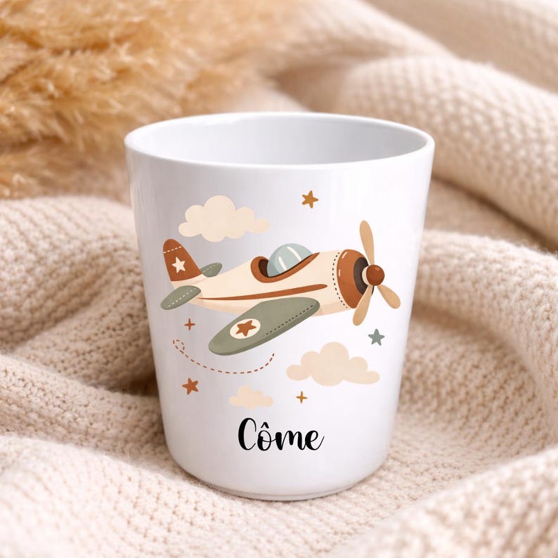 Puede incluir: Taza de cer&aacute;mica blanca con un dise&ntilde;o de avi&oacute;n de dibujos animados en marr&oacute;n, crema y verde oliva. El dise&ntilde;o incluye nubes, estrellas y el nombre "C&ocirc;me" en escritura negra. La taza es cil&iacute;ndrica y parece vac&iacute;a.