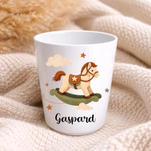 Puede incluir: Taza de cer&aacute;mica blanca con un dise&ntilde;o de caballo balanc&iacute;n de dibujos animados, nubes y estrellas. La taza tiene el nombre "Gaspard" escrito en negro. El caballo balanc&iacute;n es de color beige, marr&oacute;n y verde.