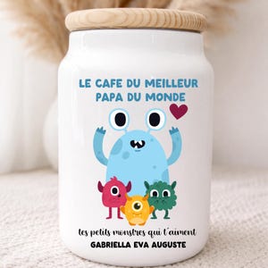 Peut inclure: Pot en céramique blanc avec un couvercle en bois, orné d'une illustration de monstre bleu avec trois petits monstres. Le texte en français indique "LE CAFE DU MEILLEUR PAPA DU MONDE" et "tes petits monstres qui t'aiment" avec un nom en dessous.