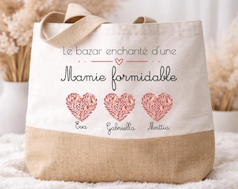Puede incluir: Un bolso tote de lona con base y asas beige. El bolso muestra el texto "Le bazar enchanté d'une Mamie formidable" y tres diseños en forma de corazón con los nombres Eva, Gabriella y Mattia.