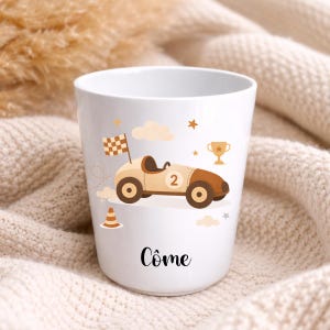 Puede incluir: Taza de cer&aacute;mica blanca con un dise&ntilde;o de coche de carreras de dibujos animados. El coche es beige y marr&oacute;n, con una bandera a cuadros, el n&uacute;mero 2 y el nombre "C&ocirc;me". Incluye estrellas y nubes, un trofeo y un cono de tr&aacute;fico. Apta para bebidas fr&iacute;as o calientes.