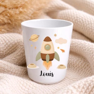 Puede incluir: Taza de cer&aacute;mica blanca con un cohete de dibujos animados, planetas, estrellas y nubes. El cohete es marr&oacute;n, beige y verde, con llamas naranjas. El nombre "Louis" est&aacute; escrito en escritura negra en la parte inferior de la taza.