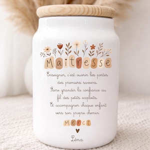 Puede incluir: Un tarro de cerámica blanco con tapa de madera, decorado con detalles florales y la palabra "Maitresse". El tarro presenta un mensaje sincero en francés, con la palabra "Merci" y un nombre en la parte inferior. Un regalo atento.