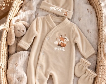Könnte beinhalten: Ein beiges Baby-Set bestehend aus einem langärmeligen Strampler, einer Mütze und Fäustlingen. Der Strampler und die Mütze zeigen den Namen Gabriel in weißer Schreibschrift, einen Cartoon-Fuchs, Mond und Sterne. Ein Plüschhase ist ebenfalls enthalten.