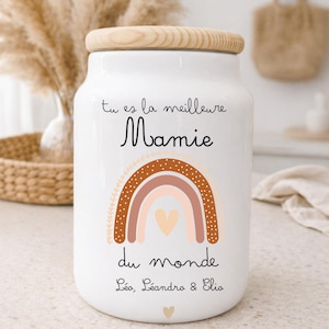 Peut inclure: Pot en céramique blanc avec un couvercle en bois, portant l'inscription "tu es la meilleure Mamie du monde" et des noms. Un motif arc-en-ciel brun, pêche et crème est en dessous. Un cœur est en bas.