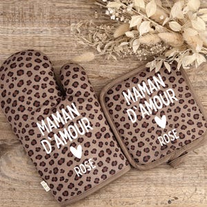May include: A brown oven mitt and pot holder set with a leopard print pattern. The text "MAMAN D'AMOUR ROSE" is printed in white, along with a white heart. The set is made of a soft, textured fabric.