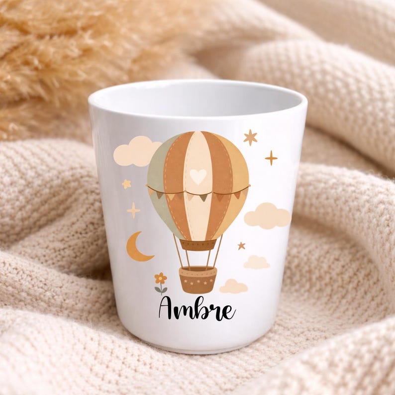 Puede incluir: Taza de cer&aacute;mica blanca con un dise&ntilde;o de globo aerost&aacute;tico en tonos marr&oacute;n, beige y verde, con nubes, estrellas y una luna creciente. El nombre "Ambre" est&aacute; escrito debajo. Un regalo encantador.