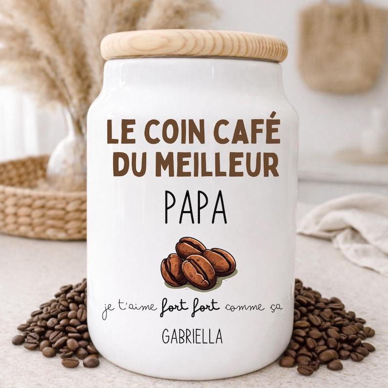 May include: A white ceramic coffee jar with a wooden lid. The jar has the text "LE COIN CAF&Eacute; DU MEILLEUR PAPA" and "je t'aime fort fort comme &ccedil;a GABRIELLA" with a graphic of coffee beans. Coffee beans surround the jar.