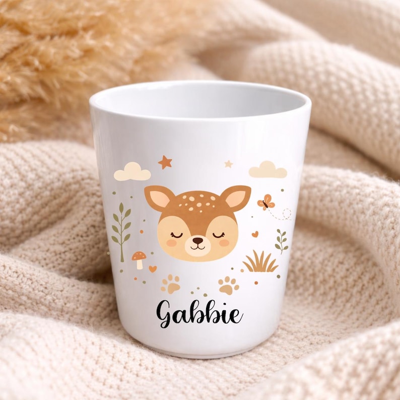 Pu&ograve; includere: Bicchiere in ceramica bianca con un design a cervo dei cartoni animati. Il cervo ha macchie marroni, occhi chiusi e un'espressione sorridente. Decorato con nuvole, stelle e il nome "Gabbie" in corsivo. Include impronte di zampe, un fungo e fogliame.