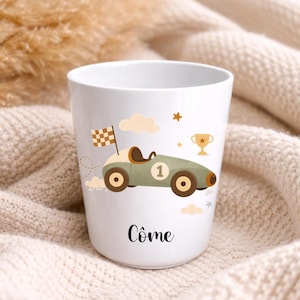 Puede incluir: Taza de cer&aacute;mica blanca con un dise&ntilde;o de coche de carreras de dibujos animados en verde oliva, marr&oacute;n y beige, con el n&uacute;mero 1. El dise&ntilde;o incluye una bandera a cuadros, nubes, estrellas y un trofeo. El nombre "C&ocirc;me" est&aacute; impreso en escritura negra en la parte inferior.