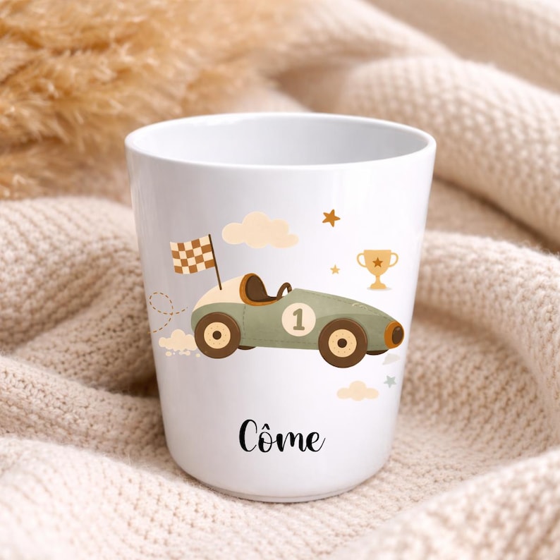 Puede incluir: Taza de cer&aacute;mica blanca con un dise&ntilde;o de coche de carreras de dibujos animados en verde oliva, marr&oacute;n y beige, con el n&uacute;mero 1. El dise&ntilde;o incluye una bandera a cuadros, nubes, estrellas y un trofeo. El nombre "C&ocirc;me" est&aacute; impreso en escritura negra en la parte inferior.