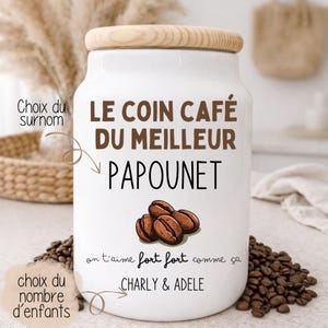 May include: A white ceramic coffee jar with a wooden lid. The jar features the text "LE COIN CAF&Eacute; DU MEILLEUR PAPOUNET" in brown, along with a graphic of coffee beans. The text "on t'aime fort fort comme &ccedil;a CHARLY & ADELE" is also present.