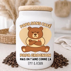 Peut inclure: Pot en céramique blanc avec un couvercle en bois, orné d'une illustration d'ours de dessin animé sur un fond en forme de cœur. Le texte dit "PAPA SANS CAFÉ" et "PAPA GROGNON MAIS ON T'AIME COMME ÇA EMY & ROBIN". Des grains de café entourent le pot.