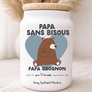 Può includere: Un barattolo di ceramica bianca con coperchio in legno. Il barattolo presenta la scritta "PAPA SANS BISOUS" sopra un'illustrazione di un orso marrone all'interno di un cuore blu. Sotto l'orso c'è la scritta "PAPA GROGNON" e la frase "mais on t'aime comme ça".