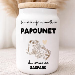 Puede incluir: Un tarro de café de cerámica blanca con tapa de madera. El tarro tiene el texto "le pot à café du meilleur PAPOUNET du monde GASPARD" en negro, junto con un dibujo de dos cápsulas de café y un corazón.