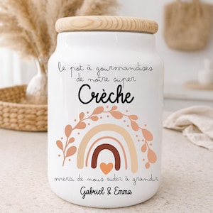 Peut inclure: Pot en céramique blanc avec un couvercle en bois. Le pot présente un motif arc-en-ciel dans les tons marron et pêche, avec les mots "Crèche" et d'autres textes en français. C'est un objet décoratif.