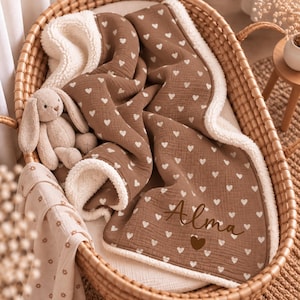 Peut inclure: Une couverture pour bébé marron avec un motif de cœurs blancs et une doublure en sherpa blanc, portant le prénom Alma brodé en écriture cursive marron foncé au-dessus d'un cœur marron plein. Un petit lapin en peluche beige est posé dessus.
