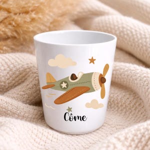 Puede incluir: Taza de cer&aacute;mica blanca con un dise&ntilde;o de avi&oacute;n de dibujos animados en verde y marr&oacute;n, con estrellas y nubes. La taza tiene el nombre "C&ocirc;me" en escritura negra. La taza est&aacute; sobre un fondo neutro.