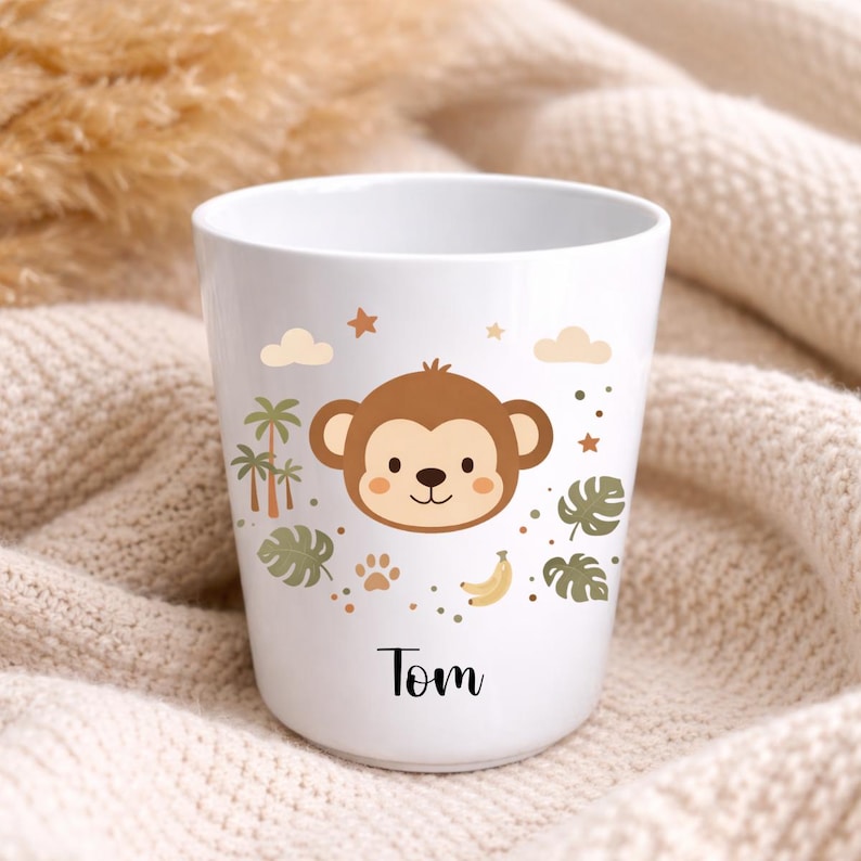 Pu&ograve; includere: Una tazza in ceramica bianca con un'illustrazione di una faccia di scimmia marrone, palme, foglie di monstera, banane, nuvole e stelle. Il nome Tom &egrave; stampato in corsivo nero sotto il disegno della scimmia sulla parte frontale.