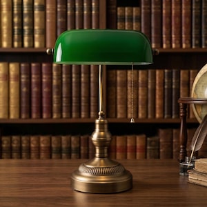 Puede incluir: Una lámpara de escritorio de estilo vintage con una pantalla de vidrio verde y una base de latón. La lámpara está colocada sobre un escritorio de madera frente a una estantería llena de libros antiguos. Un globo terráqueo y libros también están en el escritorio.