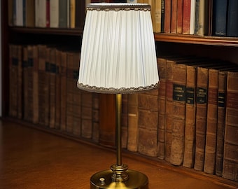 May include: A small, brass-colored table lamp with a pleated white lampshade. The lampshade is trimmed with a decorative brown border. The lamp is on a wooden surface in front of a bookshelf filled with books.