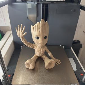 Puede incluir: Una figura de Groot, de color marrón claro, impresa en 3D, de Guardianes de la Galaxia. La figura está sentada sobre una placa de metal dentro de una impresora 3D. Groot está en una pose de saludo.