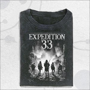 Puede incluir: Camiseta gris oscuro con las palabras "EXPEDITION 33" en blanco. La camiseta presenta un gráfico de cinco figuras con mochilas y bastones de senderismo, frente a un paisaje montañoso. La camiseta está doblada.