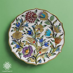 Pode incluir: Um prato de cerâmica decorativo com fundo branco e borda dourada. O prato apresenta um desenho floral com flores vermelhas, azuis, amarelas e roxas. O prato tem uma borda ondulada e as palavras "Ceramic Whisper Art" estão na parte inferior.