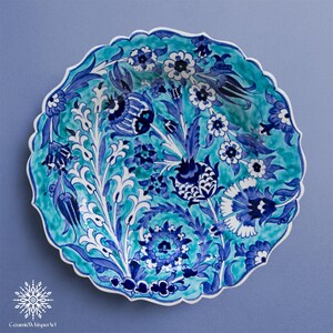 May include: A round ceramic plate with a scalloped edge, featuring a floral design in shades of blue and white against a turquoise background. The plate has a glossy finish and is likely handmade. The text "CeramicWhisper Art" is at the bottom.