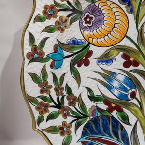 Puede incluir: Un plato decorativo de cer&aacute;mica con una superficie blanca texturizada y un borde dorado. El plato presenta un colorido dise&ntilde;o floral con flores naranjas, moradas, azules y rojas, y hojas verdes.