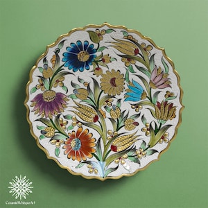 May include: A decorative ceramic plate with a white base and a scalloped gold rim. The plate features a floral design with blue, orange, yellow, and purple flowers, green leaves, and gold accents. The text "Ceramic Whisper Art" is at the bottom.