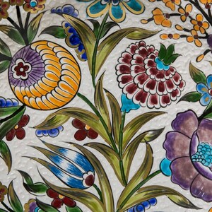 Puede incluir: Primer plano de una baldosa de cer&aacute;mica blanca con un colorido estampado floral. El dise&ntilde;o presenta flores estilizadas en tonos naranja, morado, azul y burdeos, con hojas y tallos verdes. El fondo tiene una superficie texturizada y ligeramente irregular.
