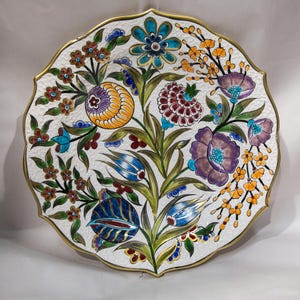 Puede incluir: Un plato decorativo de cer&aacute;mica con borde festoneado y borde dorado. El plato presenta un vibrante dise&ntilde;o floral con flores en tonos azules, morados, naranjas y marrones sobre un fondo blanco. El plato tiene una superficie texturizada.