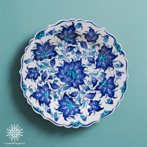 Op de afbeelding: Een ronde, witte keramische plaat met een geschulpte rand, versierd met een gedetailleerd bloemmotief in blauwe en turquoise tinten. De bloemen en bladeren zijn symmetrisch gerangschikt. De plaat heeft een getextureerd oppervlak.