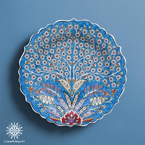 Può includere: Un piatto rotondo in ceramica blu con bordo smerlato. Il piatto presenta un motivo ad albero della vita con fiori bianchi, dettagli rossi e un motivo floreale dettagliato. Il piatto ha un bordo bianco ed è posto su uno sfondo blu.